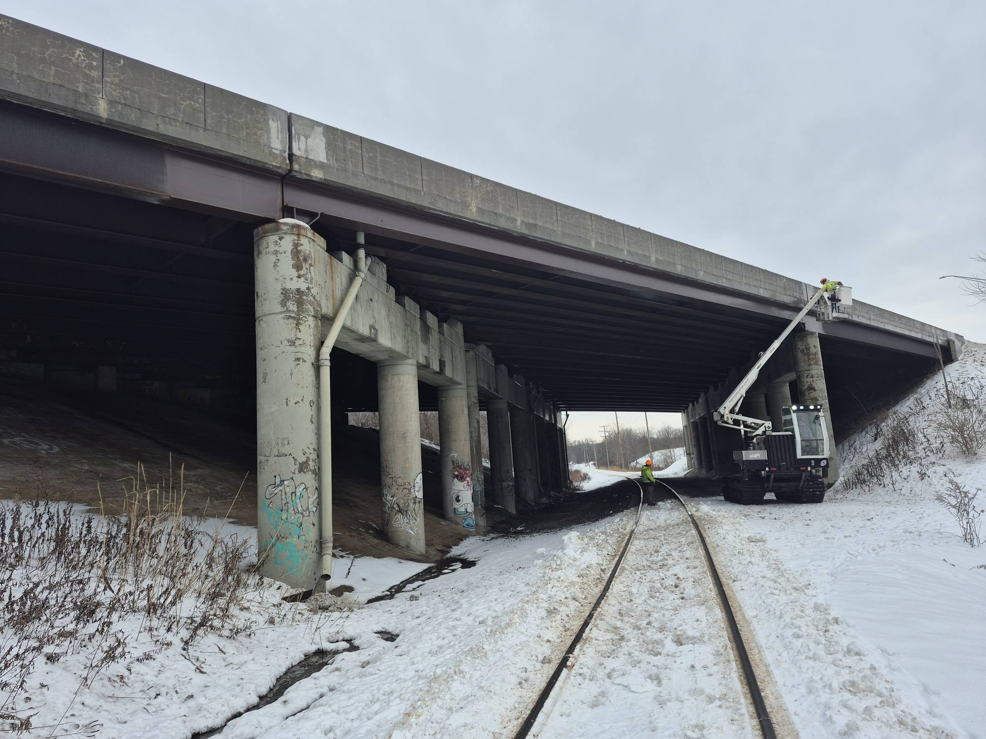 Under-bridge inspection unit reaching beneath a steel multi-girder overpass above an active rail line in winter.