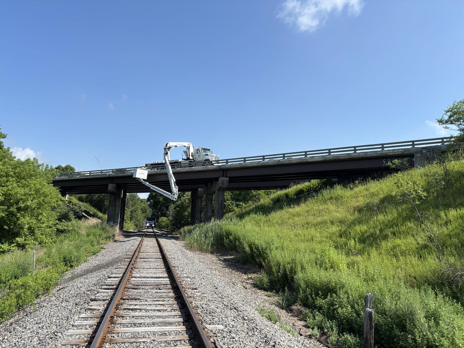 An under-bridge inspection truck positioned on a bridge deck with its boom lowered beneath the superstructure over a rail corridor.
